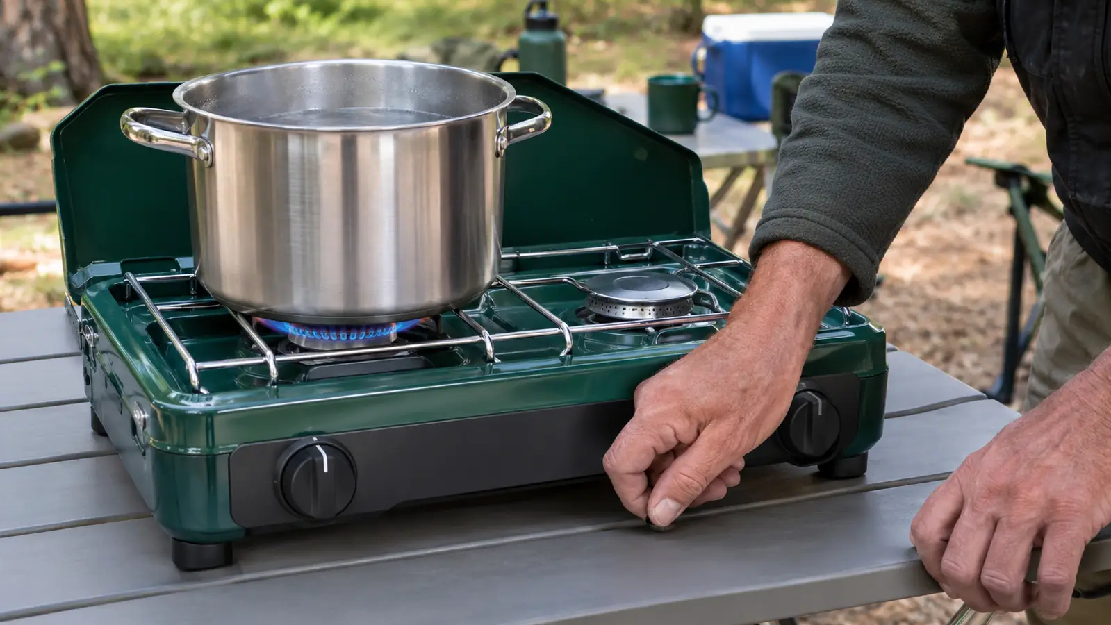 A hand adjusting the valve knob on a two-burner camp stove while a large pot of water sits on the burner grate