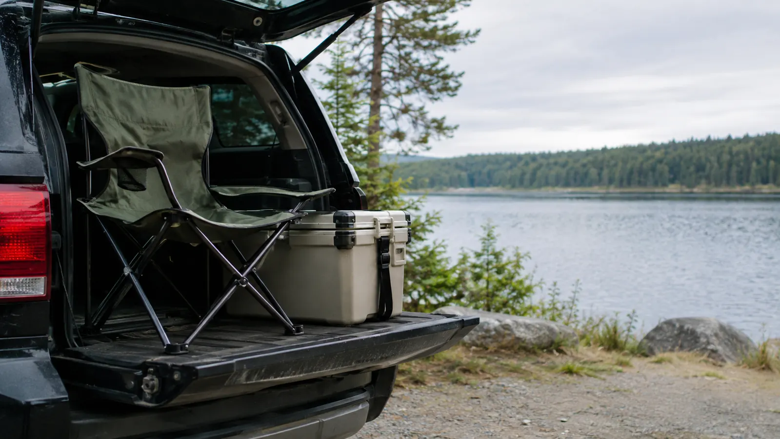 A camp chair with a cooler sits on an open tailgate at a campground, with a lake and treeline behind it