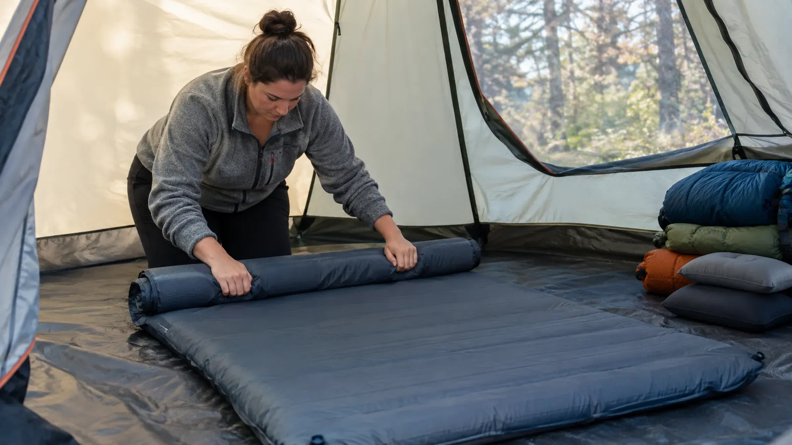 A parent rolling up a double sleeping pad from the foot end inside a tent on a camping morning, with sleeping bags stacked nearby