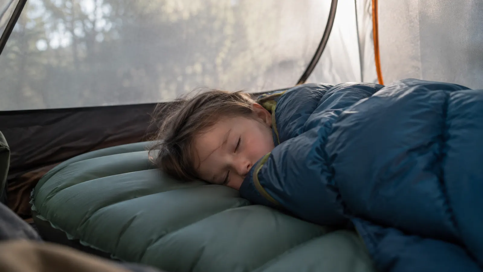 A child curled up asleep on a wide sleeping pad inside a tent, sleeping bag pulled up, soft morning light through the tent wall